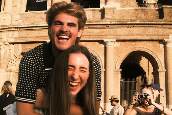A couple laughs joyfully with the Colosseum in the background, as another person takes a photo of them.