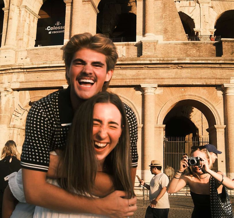 A couple laughs joyfully with the Colosseum in the background, as another person takes a photo of them.