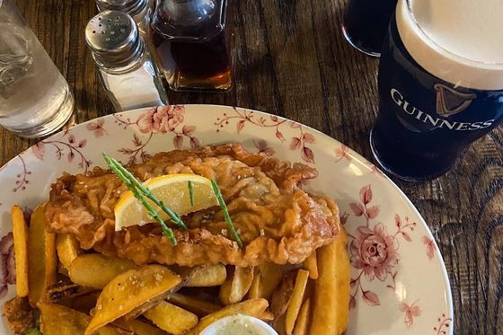 Fish and chips with mushy peas and tartar sauce on a floral plate, accompanied by two pints of Guinness on a wooden table.