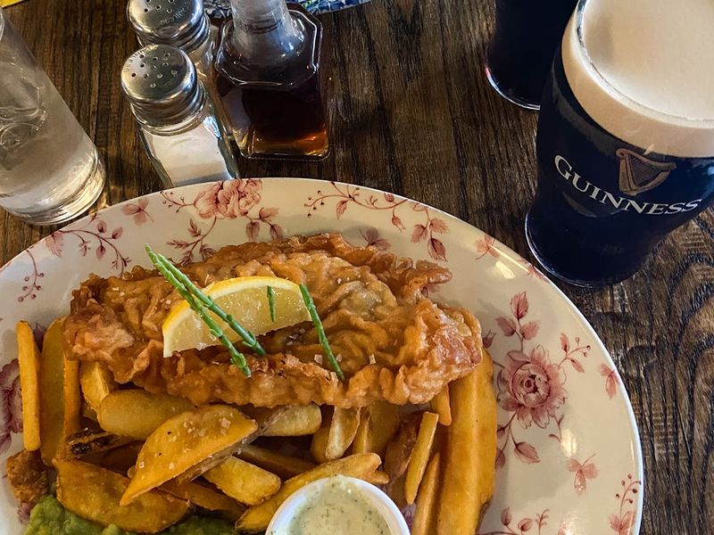 Fish and chips with mushy peas and tartar sauce on a floral plate, accompanied by two pints of Guinness on a wooden table.