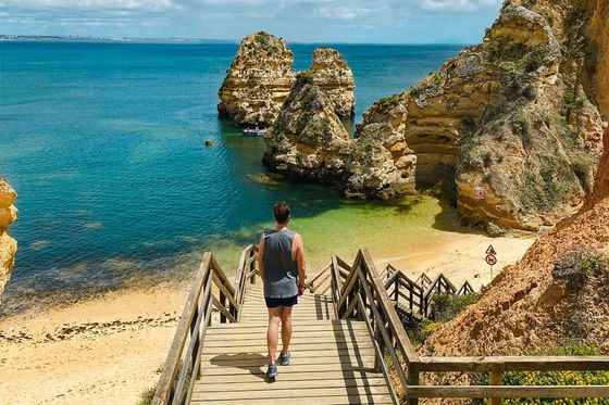 Person walking down wooden stairs to a sandy beach with rock formations and clear blue water under a partly cloudy sky.
