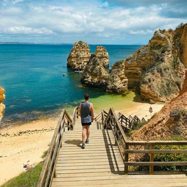 Person walking down wooden stairs to a sandy beach with rock formations and clear blue water under a partly cloudy sky.