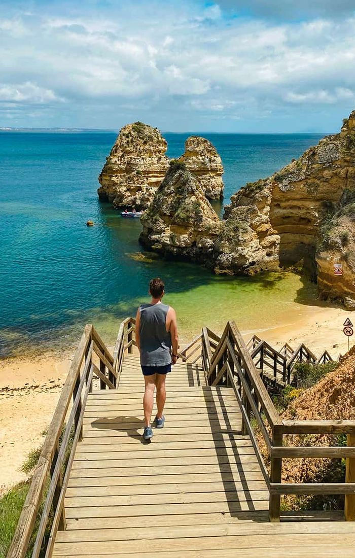 Person walking down wooden stairs to a sandy beach with rock formations and clear blue water under a partly cloudy sky.