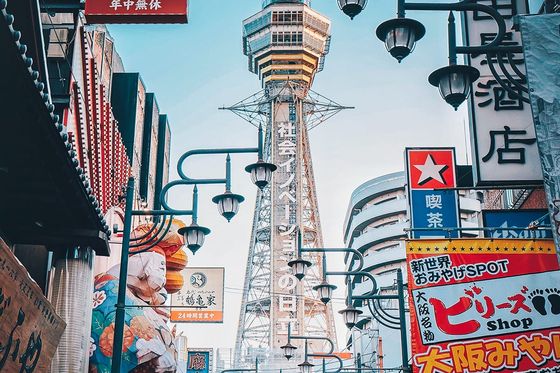 Bustling street in Osaka, Japan, featuring colorful signs, lanterns, and Tsutenkaku Tower under a clear blue sky.