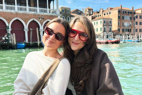 Two smiling women wearing sunglasses pose by a canal in Venice, with historic buildings and boats in the background under a sunny sky.