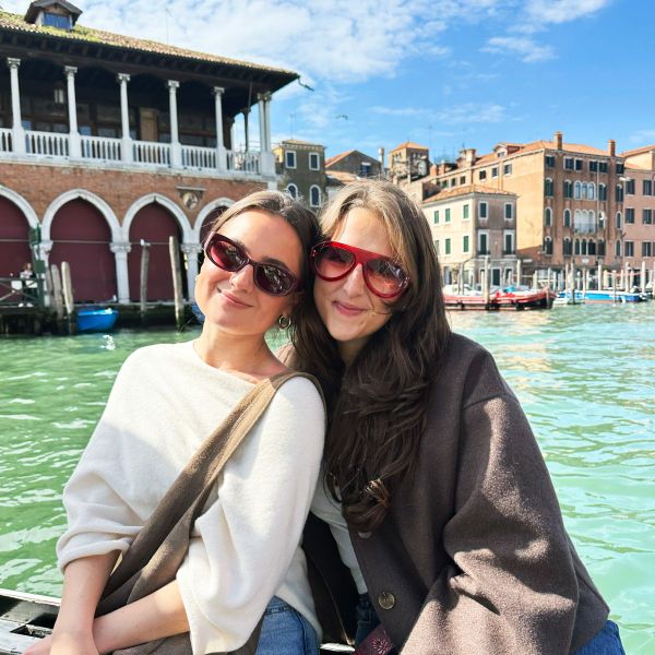 Two smiling women wearing sunglasses pose by a canal in Venice, with historic buildings and boats in the background under a sunny sky.