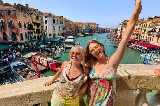 Two women smiling and posing on a bridge overlooking a Venice canal, with vibrant buildings and boats in the background on a sunny day.