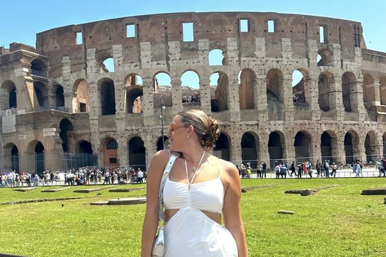 A woman in a white dress stands in front of the Colosseum in Rome, looking to the side on a sunny day.