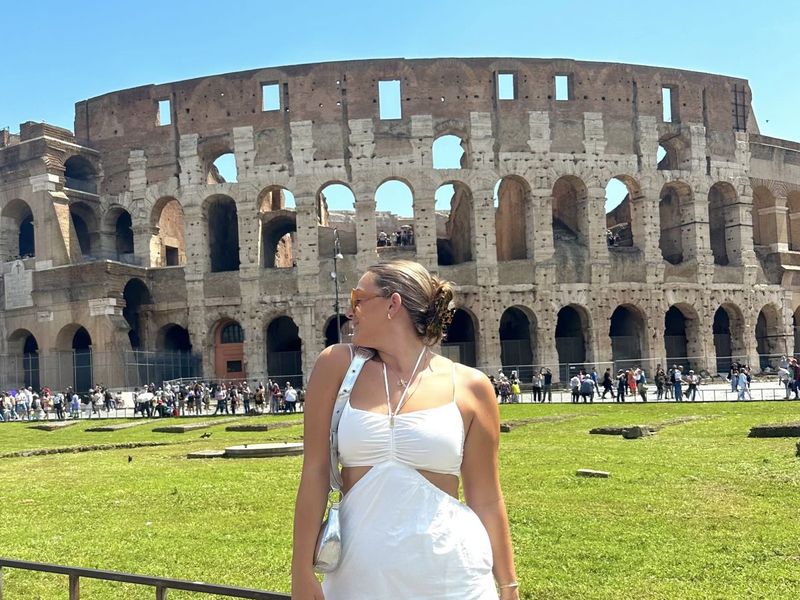 A woman in a white dress stands in front of the Colosseum in Rome, looking to the side on a sunny day.