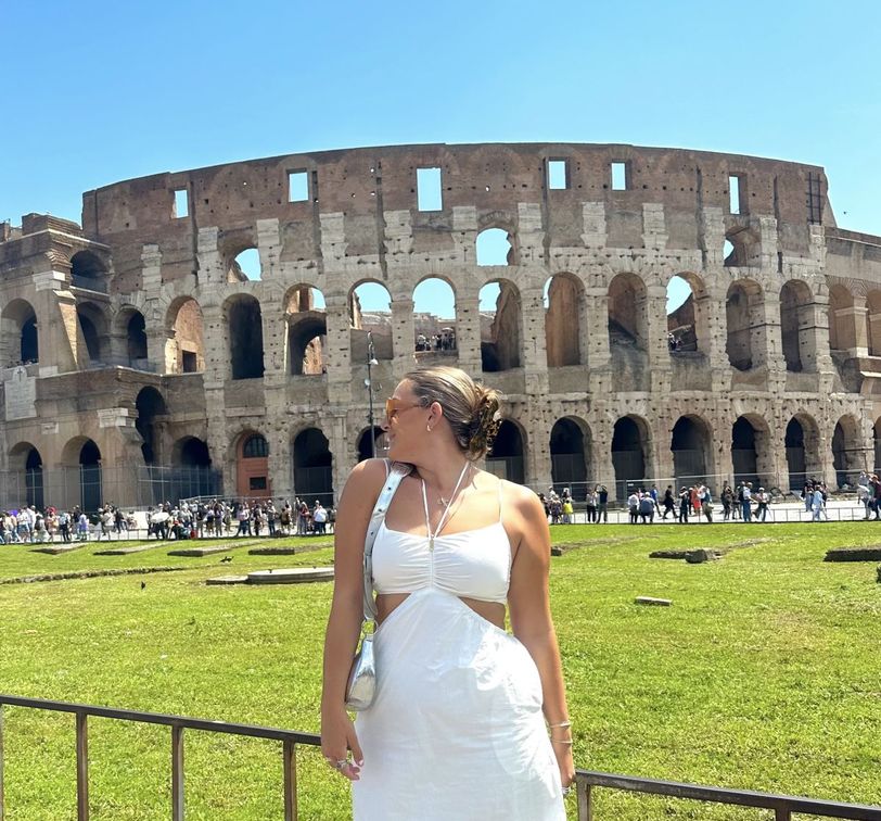 A woman in a white dress stands in front of the Colosseum in Rome, looking to the side on a sunny day.
