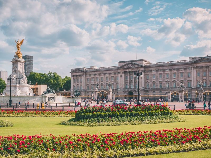 Buckingham Palace with vibrant gardens and the Victoria Memorial in the foreground, under a blue sky with fluffy clouds.