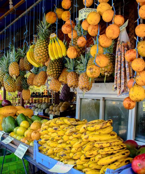 A vibrant fruit market stall with pineapples, bananas, and oranges hanging above, and various tropical fruits like mangoes and avocados displayed below.