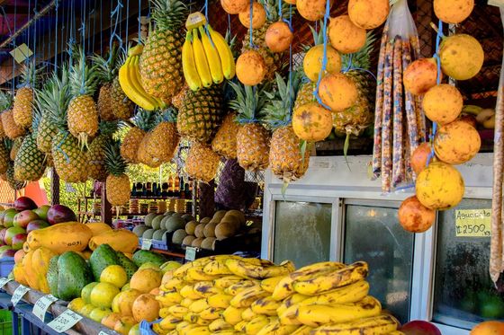 A vibrant fruit market stall with pineapples, bananas, and oranges hanging above, and various tropical fruits like mangoes and avocados displayed below.