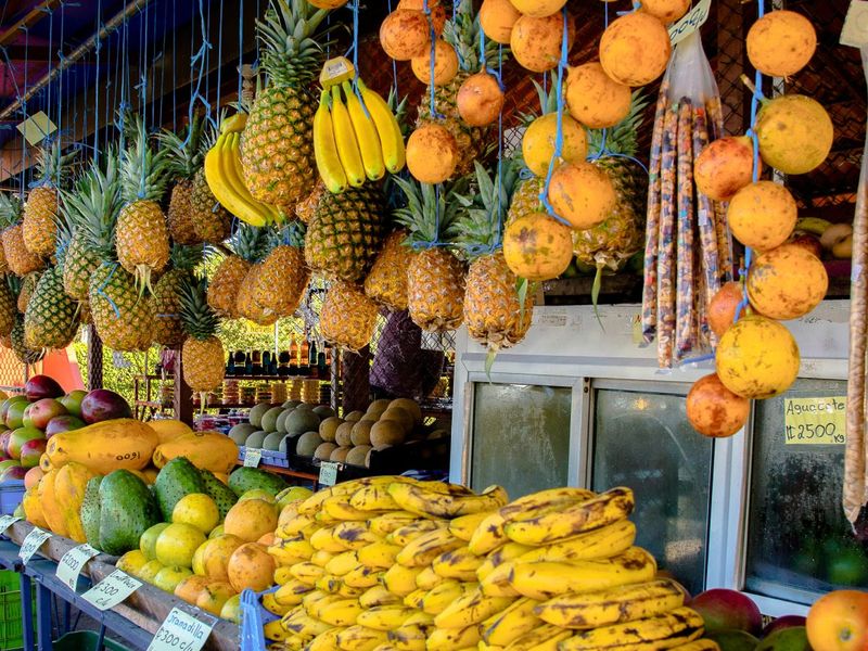 A vibrant fruit market stall with pineapples, bananas, and oranges hanging above, and various tropical fruits like mangoes and avocados displayed below.