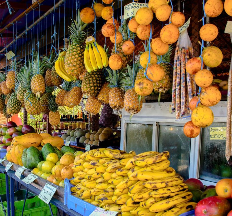 A vibrant fruit market stall with pineapples, bananas, and oranges hanging above, and various tropical fruits like mangoes and avocados displayed below.