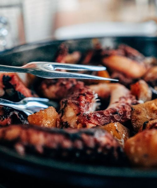 Close-up of cooked octopus and potatoes in a skillet, with metal tongs resting atop the dish.