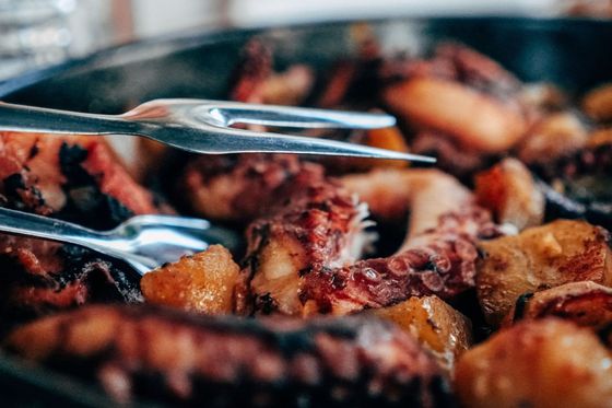 Close-up of cooked octopus and potatoes in a skillet, with metal tongs resting atop the dish.