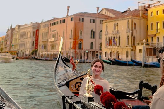 A woman smiles while sitting in a gondola on a Venetian canal, with colorful historic buildings lining the waterway in the background.
