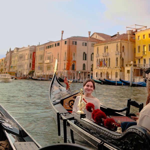 A woman smiles while sitting in a gondola on a Venetian canal, with colorful historic buildings lining the waterway in the background.