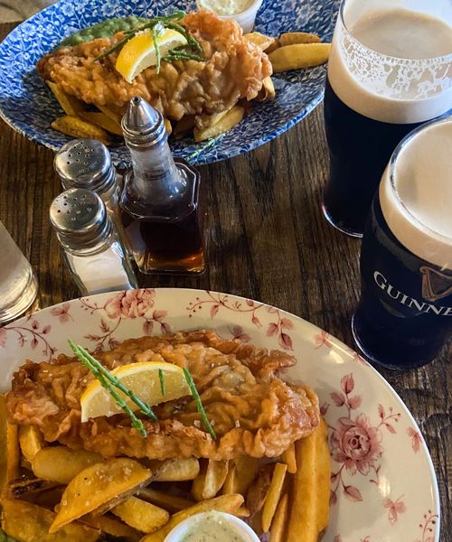 Two plates of fish and chips with lemon garnishes on a table, accompanied by pints of Guinness and condiments.