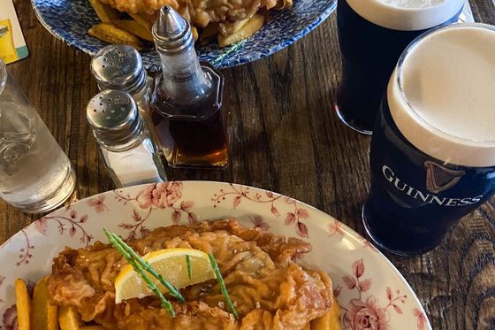 Two plates of fish and chips with lemon garnishes on a table, accompanied by pints of Guinness and condiments.