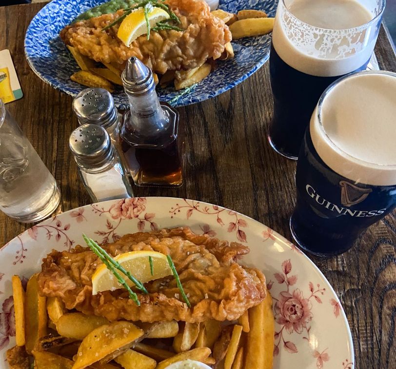 Two plates of fish and chips with lemon garnishes on a table, accompanied by pints of Guinness and condiments.