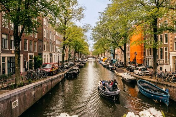 A canal flanked by trees and traditional buildings. Boats are moored along the sides, and a small boat with people is cruising on the water.