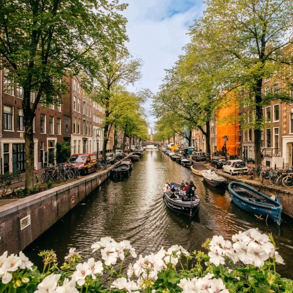 A canal flanked by trees and traditional buildings. Boats are moored along the sides, and a small boat with people is cruising on the water.