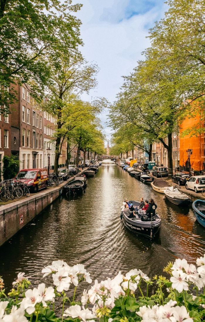 A canal flanked by trees and traditional buildings. Boats are moored along the sides, and a small boat with people is cruising on the water.
