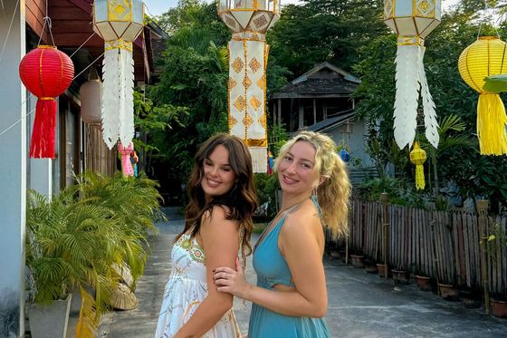 Two women in colorful dresses pose happily outdoors, surrounded by hanging paper lanterns, with trees and a wooden fence in the background.