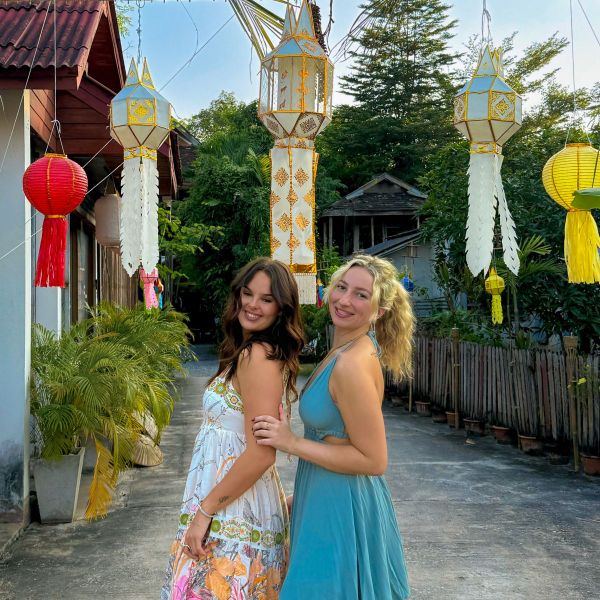 Two women in colorful dresses pose happily outdoors, surrounded by hanging paper lanterns, with trees and a wooden fence in the background.