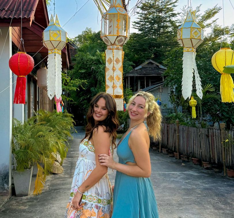 Two women in colorful dresses pose happily outdoors, surrounded by hanging paper lanterns, with trees and a wooden fence in the background.