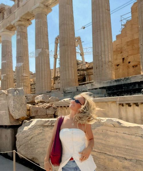 A woman in sunglasses poses in front of ancient Greek columns at the Parthenon on a sunny day, with her hair blowing in the wind.