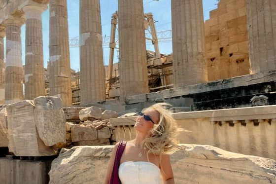 A woman in sunglasses poses in front of ancient Greek columns at the Parthenon on a sunny day, with her hair blowing in the wind.