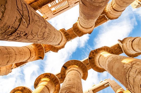 Upward view of ancient Egyptian columns with hieroglyphics, set against a bright blue sky with wispy clouds.