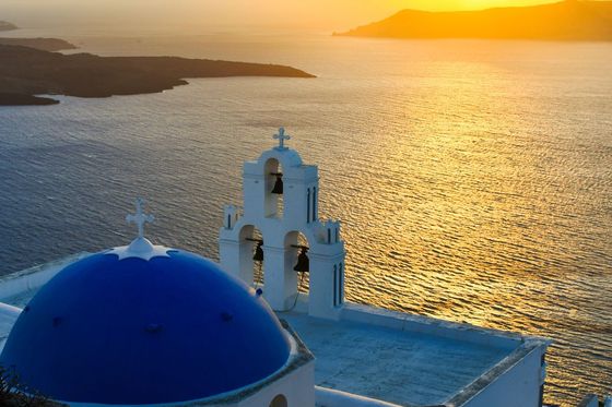 A white building with a blue dome and bell tower overlooks a golden sunset over the ocean in Santorini, Greece.