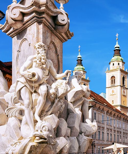 Ornate stone fountain with detailed statues, against a backdrop of historic architecture and two church towers under a clear blue sky.