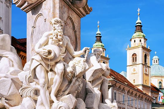 Ornate stone fountain with detailed statues, against a backdrop of historic architecture and two church towers under a clear blue sky.