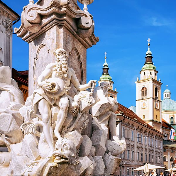 Ornate stone fountain with detailed statues, against a backdrop of historic architecture and two church towers under a clear blue sky.
