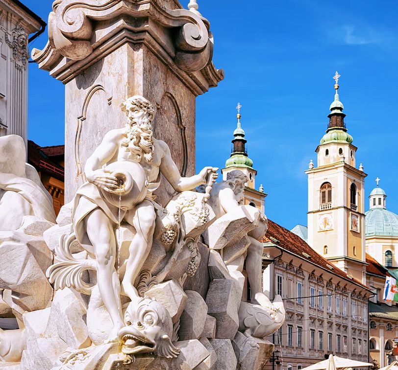 Ornate stone fountain with detailed statues, against a backdrop of historic architecture and two church towers under a clear blue sky.