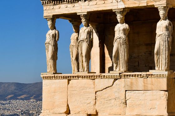Ancient Greek temple with Caryatid columns, clear blue sky, and distant cityscape in the background.
