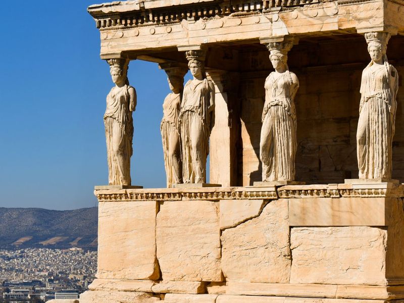 Ancient Greek temple with Caryatid columns, clear blue sky, and distant cityscape in the background.