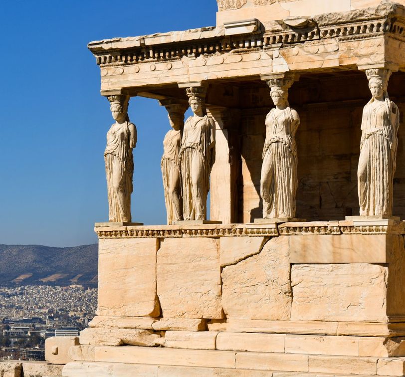 Ancient Greek temple with Caryatid columns, clear blue sky, and distant cityscape in the background.