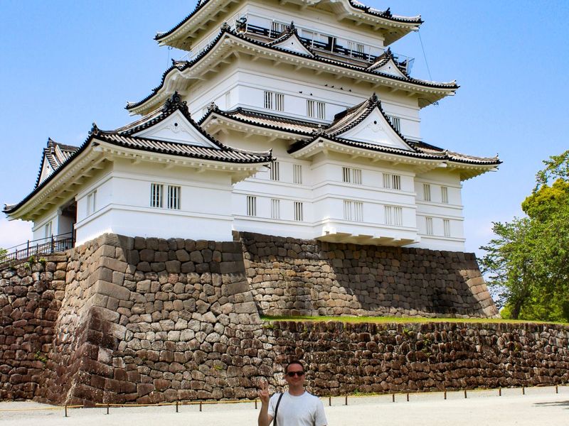 A person stands in front of a traditional Japanese castle with white walls and black tiled roofs, set on a stone foundation, under a clear blue sky.