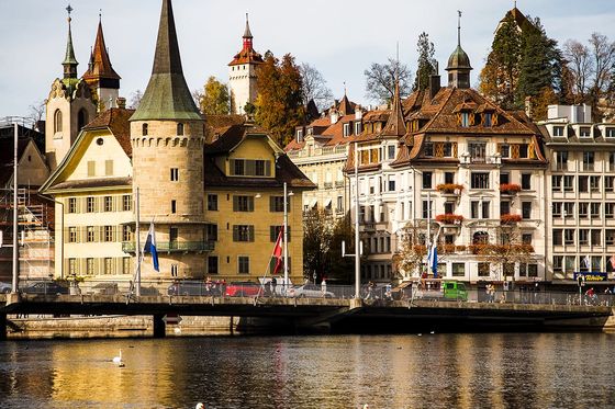 A scenic view of historic buildings and swans on a river in Lucerne, Switzerland, with a clear sky and autumn foliage.