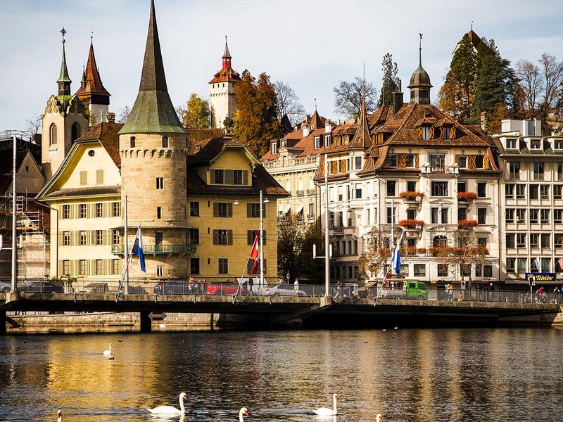 A scenic view of historic buildings and swans on a river in Lucerne, Switzerland, with a clear sky and autumn foliage.