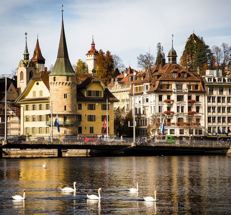 A scenic view of historic buildings and swans on a river in Lucerne, Switzerland, with a clear sky and autumn foliage.