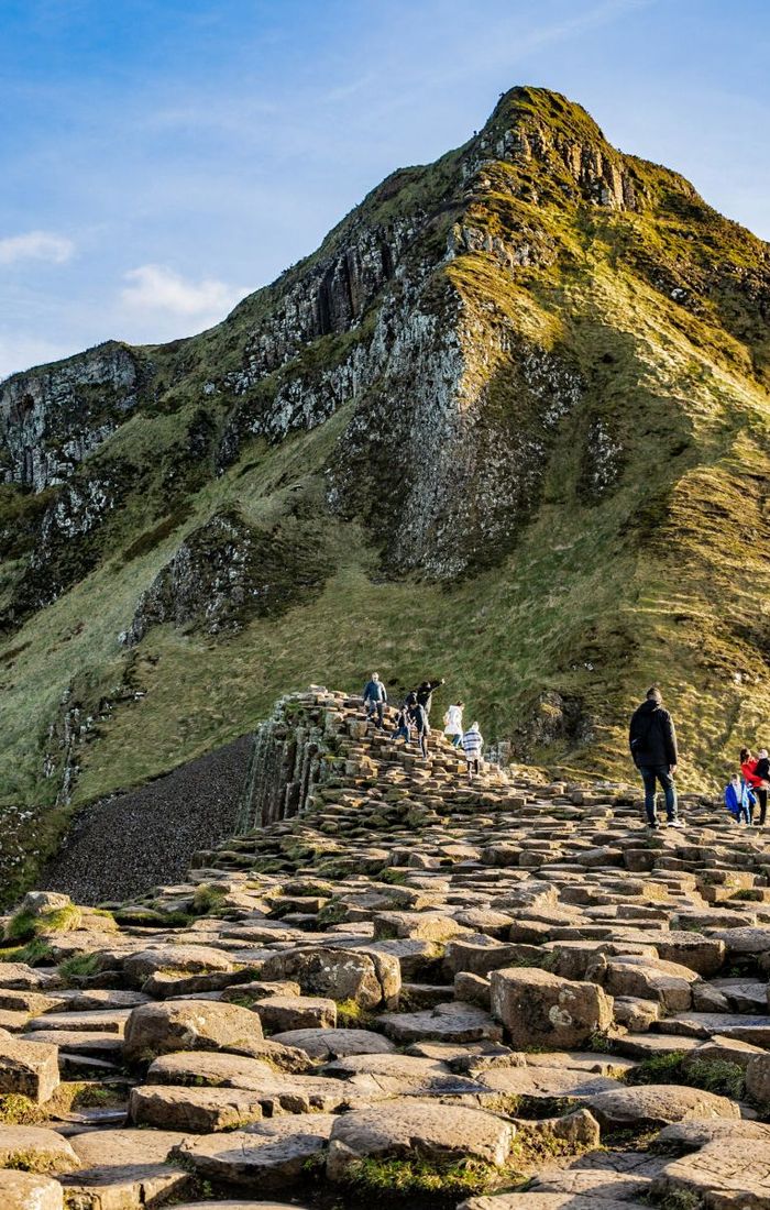 People walking on the Giant's Causeway, a natural formation of hexagonal basalt columns, with a grassy hillside and blue sky in the background.