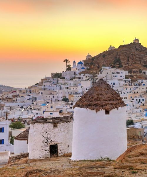 White-washed houses and an old windmill in a Greek island village at sunset, with a golden sky and a rocky hill in the background.