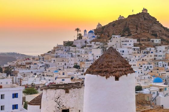 White-washed houses and an old windmill in a Greek island village at sunset, with a golden sky and a rocky hill in the background.